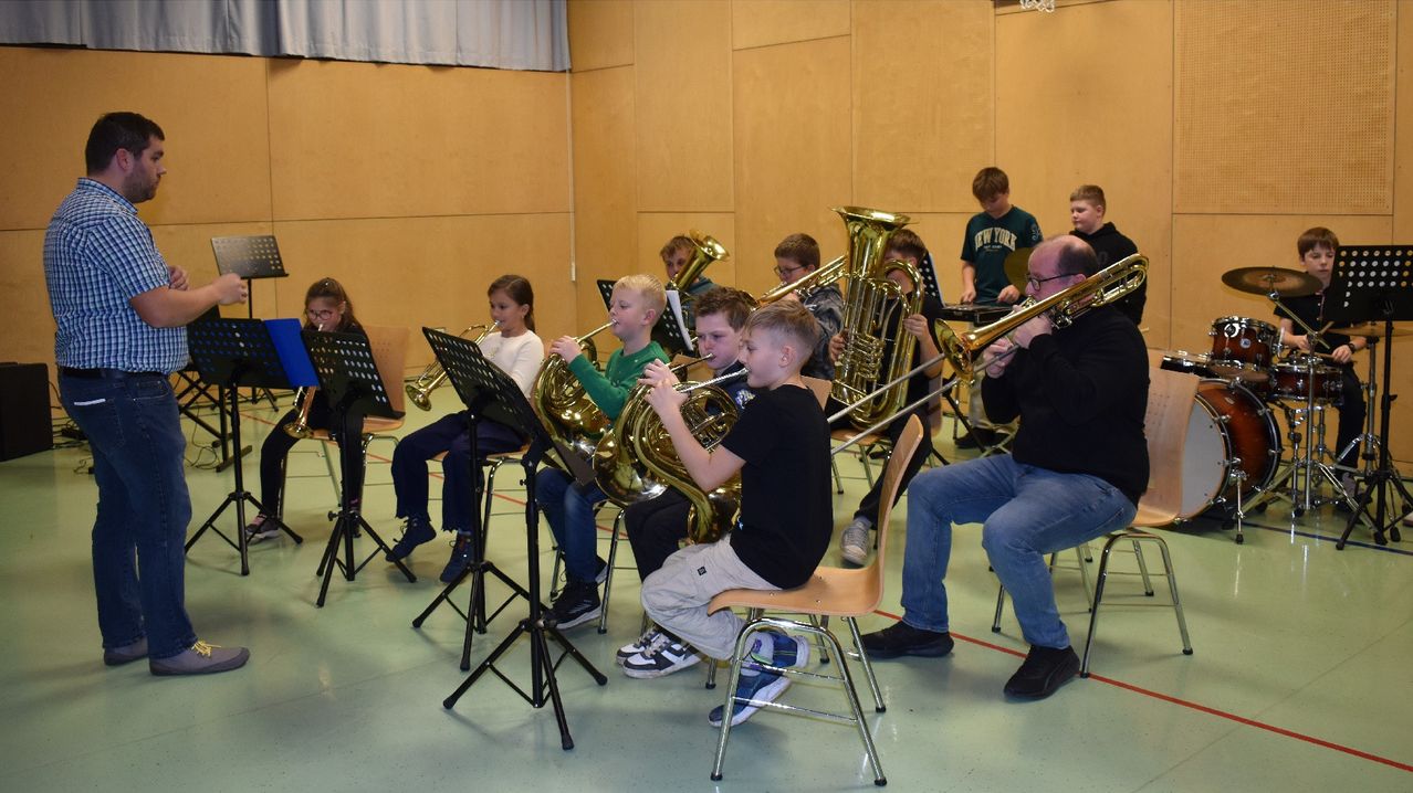 A classroom setting with children and adults playing brass instruments, seated on chairs, with music stands and a wall in the background.