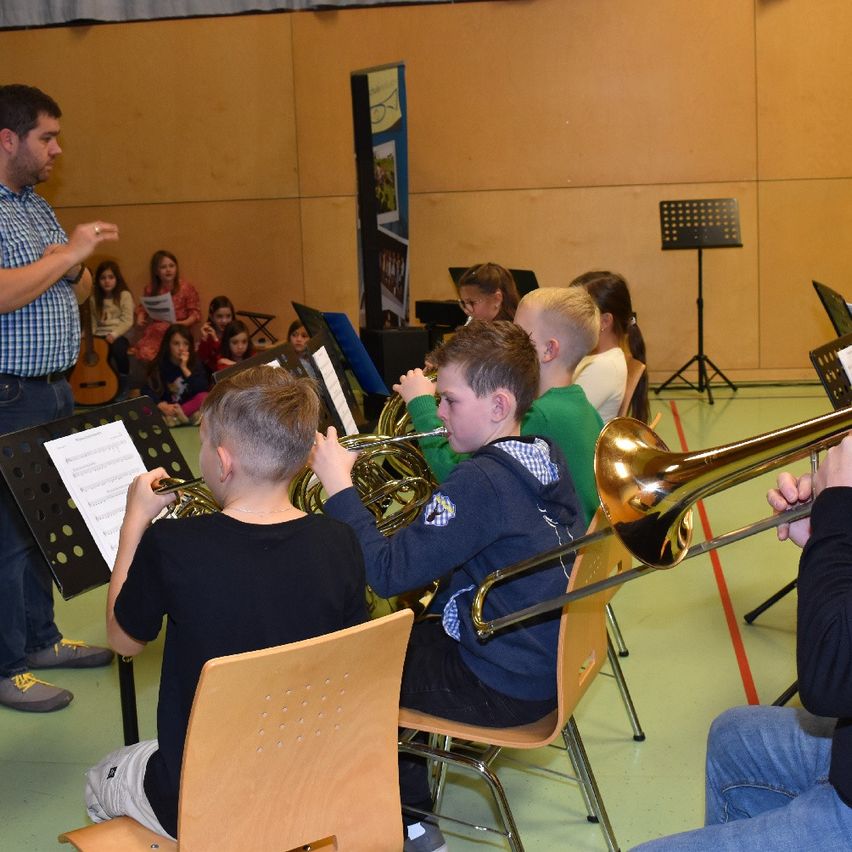A man conducts a group of children playing brass instruments. The children sit in chairs while playing. Some children stand and listen to the instructor.
