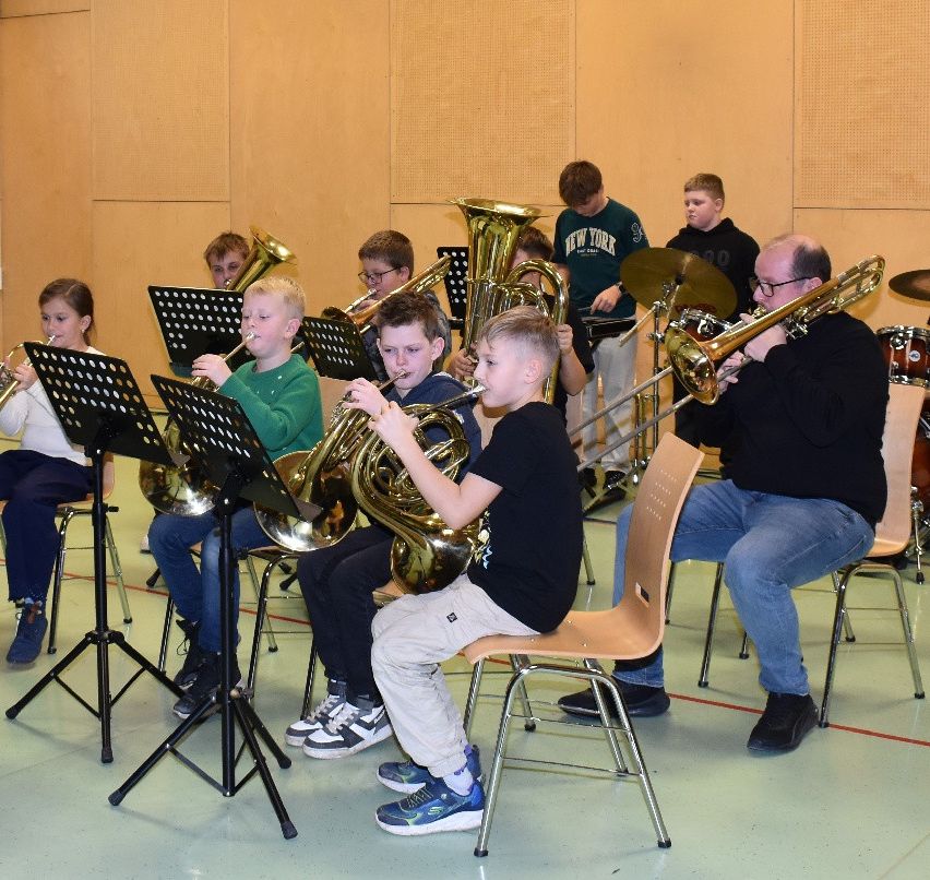 A group of children and adults are in a music room playing brass instruments, seated on chairs with music stands in front of them.