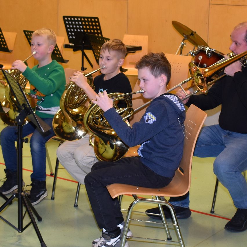 Four boys play brass instruments while seated in a classroom. One plays a trombone, two play tubas, and one plays a trumpet. An adult man watches them.