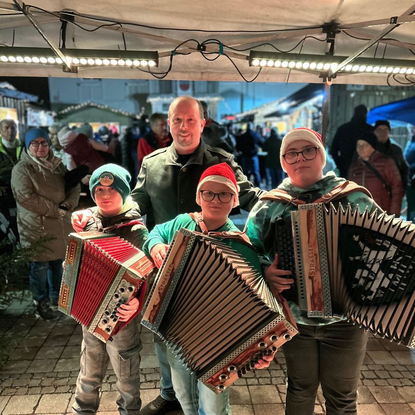 A man and three children are playing accordions in a festive setting. They are under a tent with a crowd in the background.
