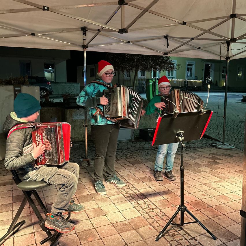 Three children in festive hats play accordions under a tent at night. The boy on the left sits on a chair while the other two stand with music stands.