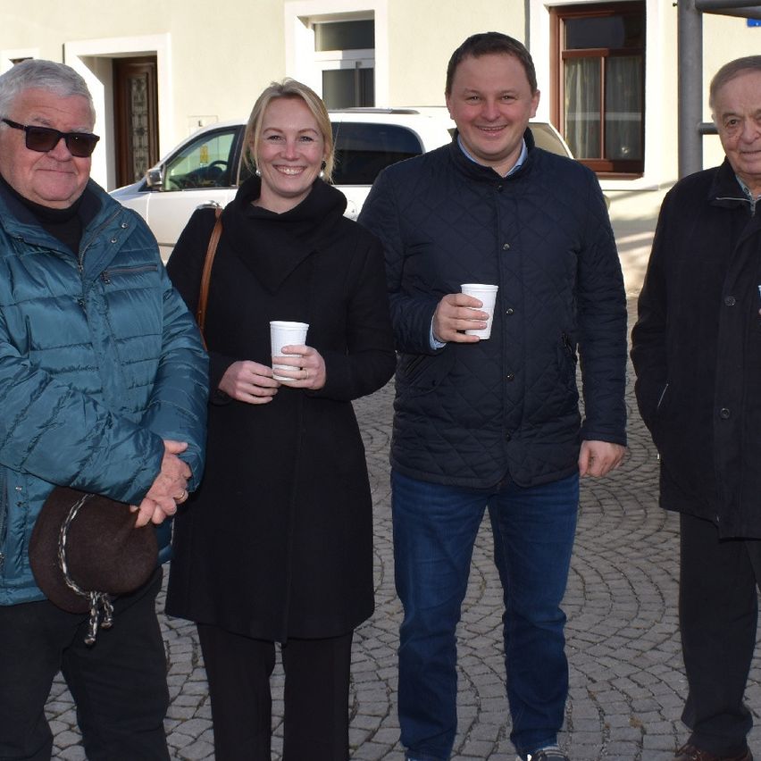 Four people are standing outside, smiling. Two are men, one with glasses, the other with a hat. Two are women, one holding a cup. Behind them is a building with windows.