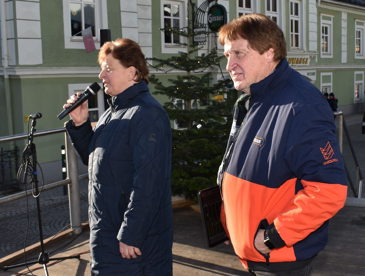 A woman with a microphone stands next to a man in front of a decorated Christmas tree. Behind them, a building with green walls and windows.
