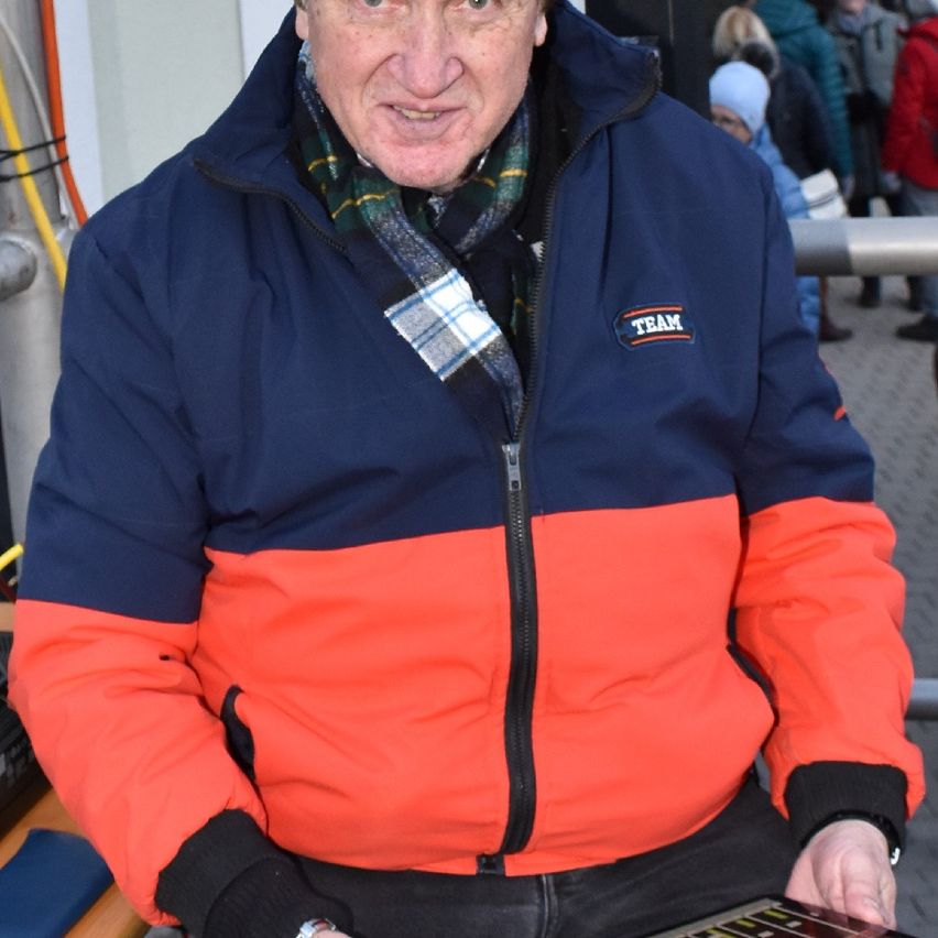 An older man in a blue and orange jacket with 'TEAM' on the left chest stands outdoors, looking at something on a device.