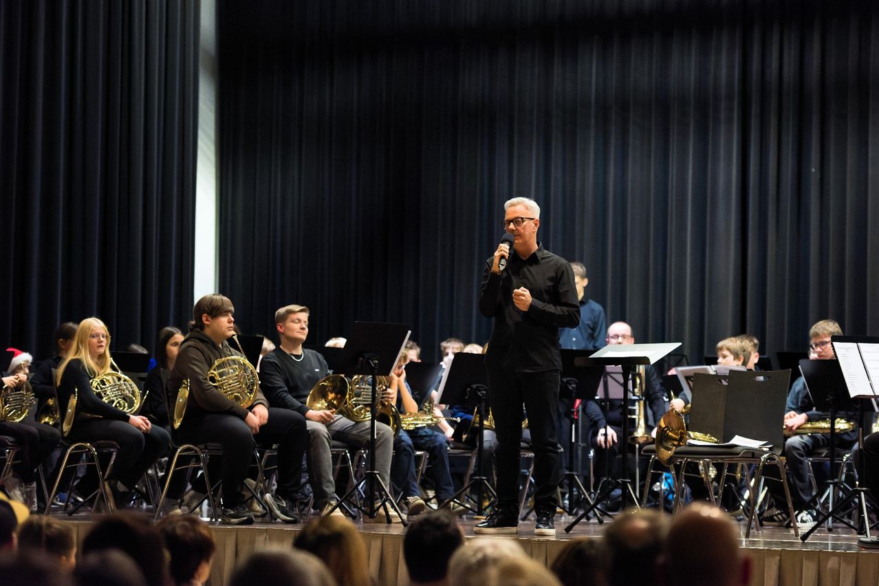 Ein Dirigent steht auf einer Bühne vor einem Orchester. Er hält ein Mikrofon und spricht das Publikum an. Die Orchestermitglieder sitzen vor ihm und halten Instrumente.