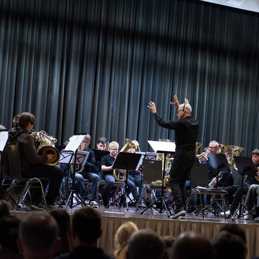 Ein Dirigent steht auf der Bühne und dirigiert mit seinen Händen ein Blasorchester, das vor einem Publikum spielt. Die Musiker sitzen auf Stühlen, und das Publikum beobachtet aufmerksam.