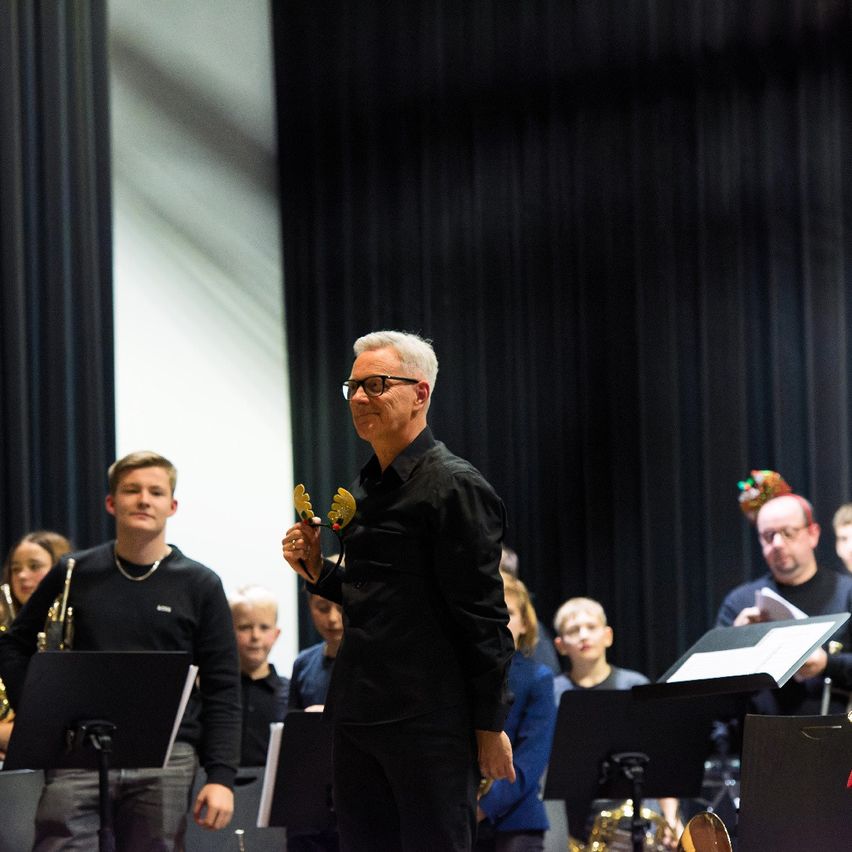 Ein Mann, der zwei Glocken hält, steht vor einer Gruppe von Musikern, wobei ein junger Mann vor ihm eine Trompete hält.