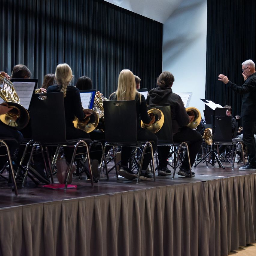 Eine Gruppe von Musikern spielt Blechblasinstrumente auf einer Bühne, angeführt von einem Dirigenten. Die Musiker sitzen auf Stühlen und die Bühne ist mit einem Vorhang bedeckt.
