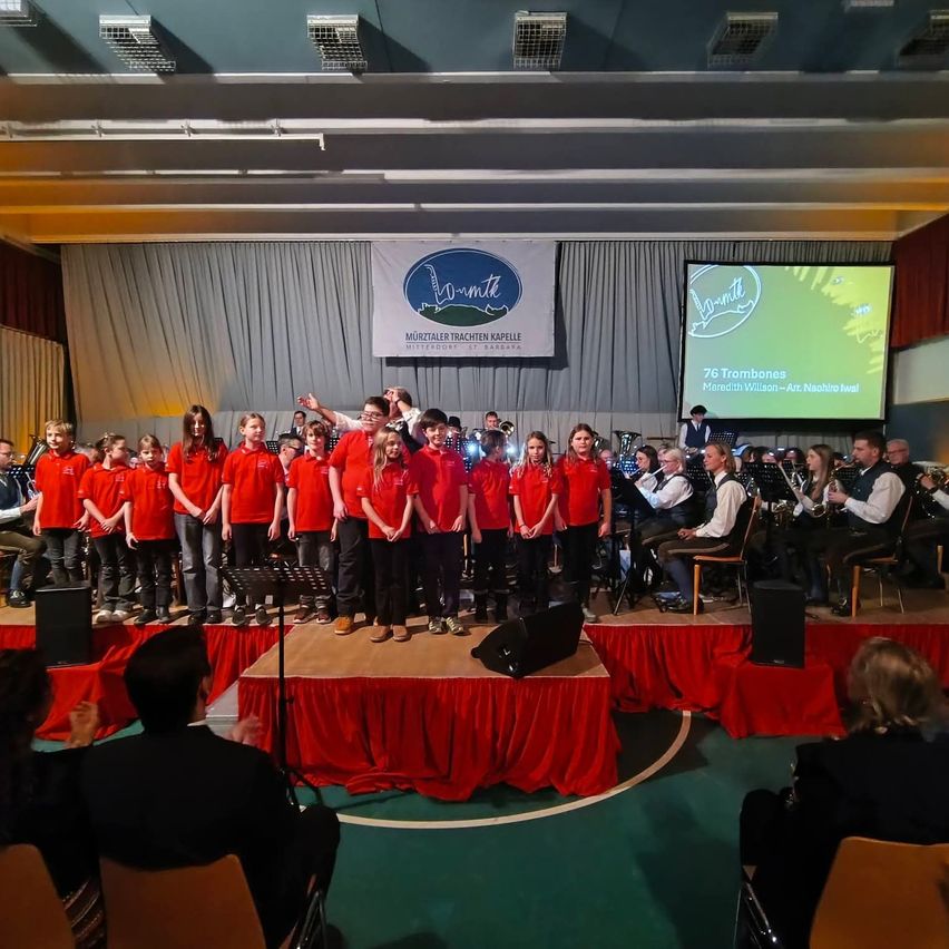 Eine Gruppe von Kindern in roten Shirts steht auf der Bühne, begleitet von einer Band, die hinter ihnen spielt. Ein großer Bildschirm mit Text und Bildern steht hinter der Band. Das Publikum sitzt auf Stühlen vor der Bühne.