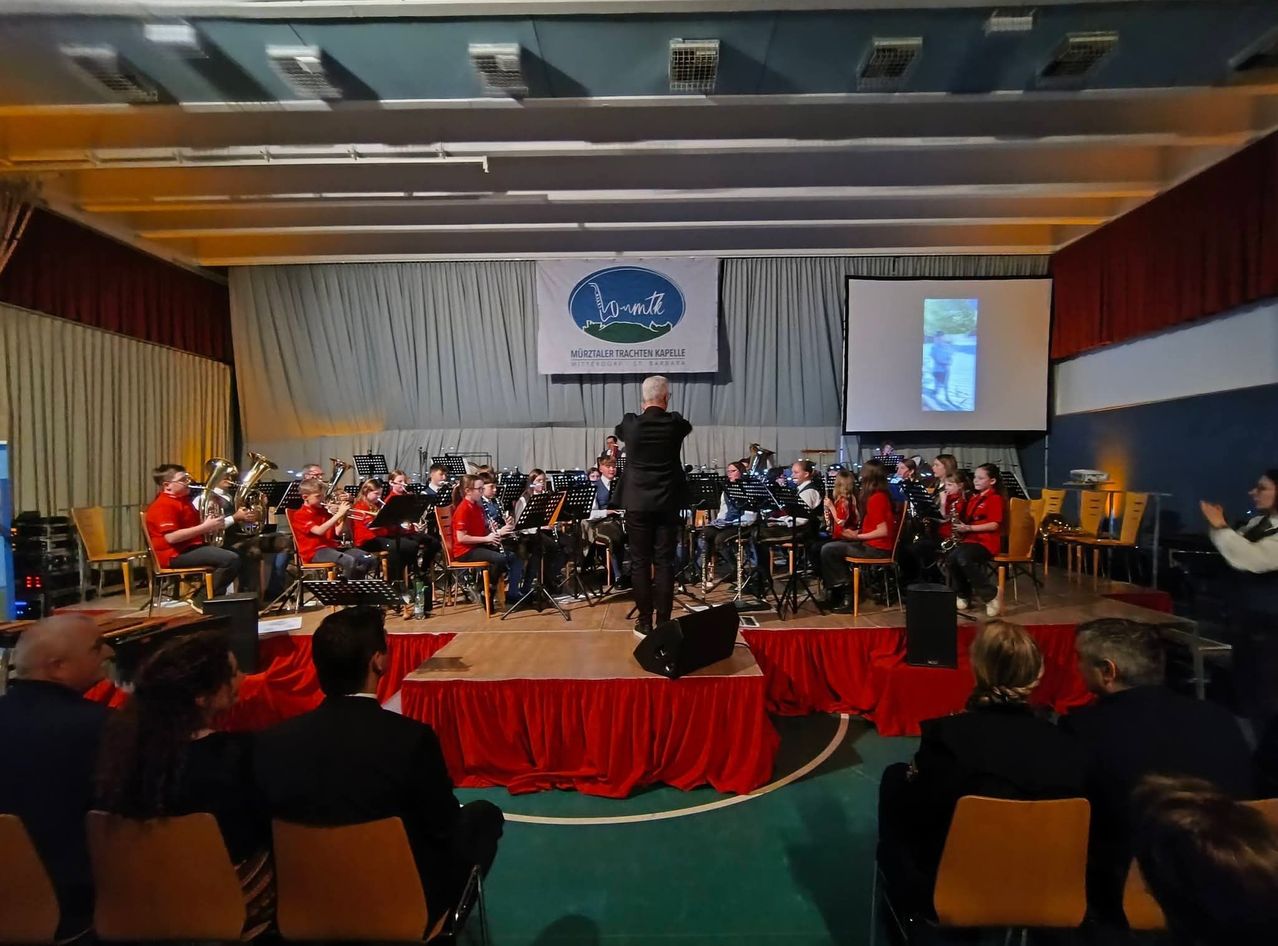 Ein Schulorchester spielt auf der Bühne, mit einem Dirigenten und verschiedenen Instrumenten. Das Publikum sitzt in Stühlen an der Seite. Ein Banner und eine Leinwand sind im Hintergrund.
