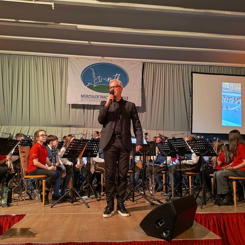 Ein Mann auf der Bühne spricht in ein Mikrofon, wahrscheinlich führt er ein Orchester oder einen Chor. Hinter ihm zeigt eine Leinwand ein Bild. Das Orchester sitzt auf Stühlen vor Notenständern.