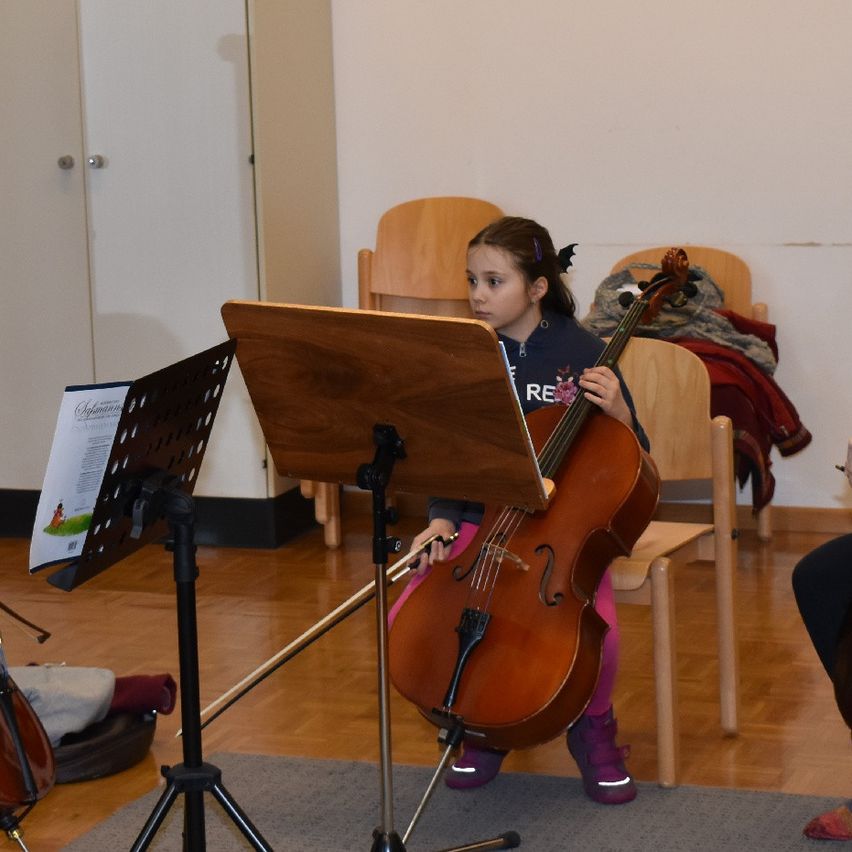 Ein kleines Mädchen spielt Cello in einem Musikraum, sitzt auf einem Stuhl mit einem Notenständer vor ihr. Dahinter stehen zwei Holzstühle und eine weiße Wand.