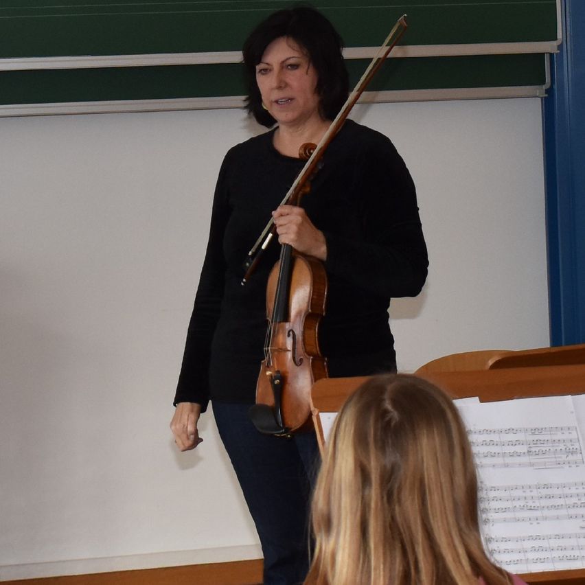 Eine Frau hält eine Geige in einem Klassenzimmer, vor ihr sitzt ein junges Mädchen und schaut sich Noten an.