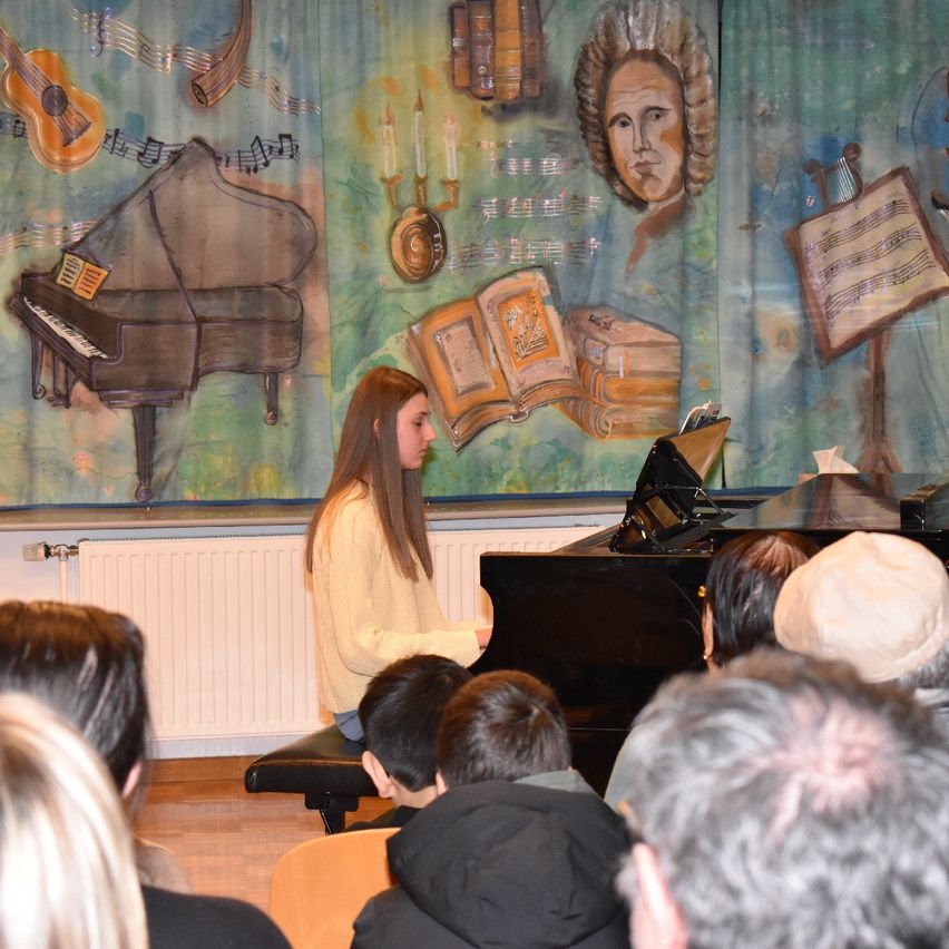 Eine Frau spielt Klavier vor einem Publikum. Hinter ihr befindet sich ein Wandbild mit einem Klavier, einem Porträt und Musiknoten. Das Publikum sitzt vor ihr.