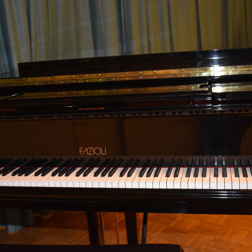 Ein schwarzer Fazioli-Flügel mit goldenen Akzenten steht in einem Raum mit Holzboden. Der Flügel hat eine glänzende Oberfläche und eine schwarz-weiße Tastatur.