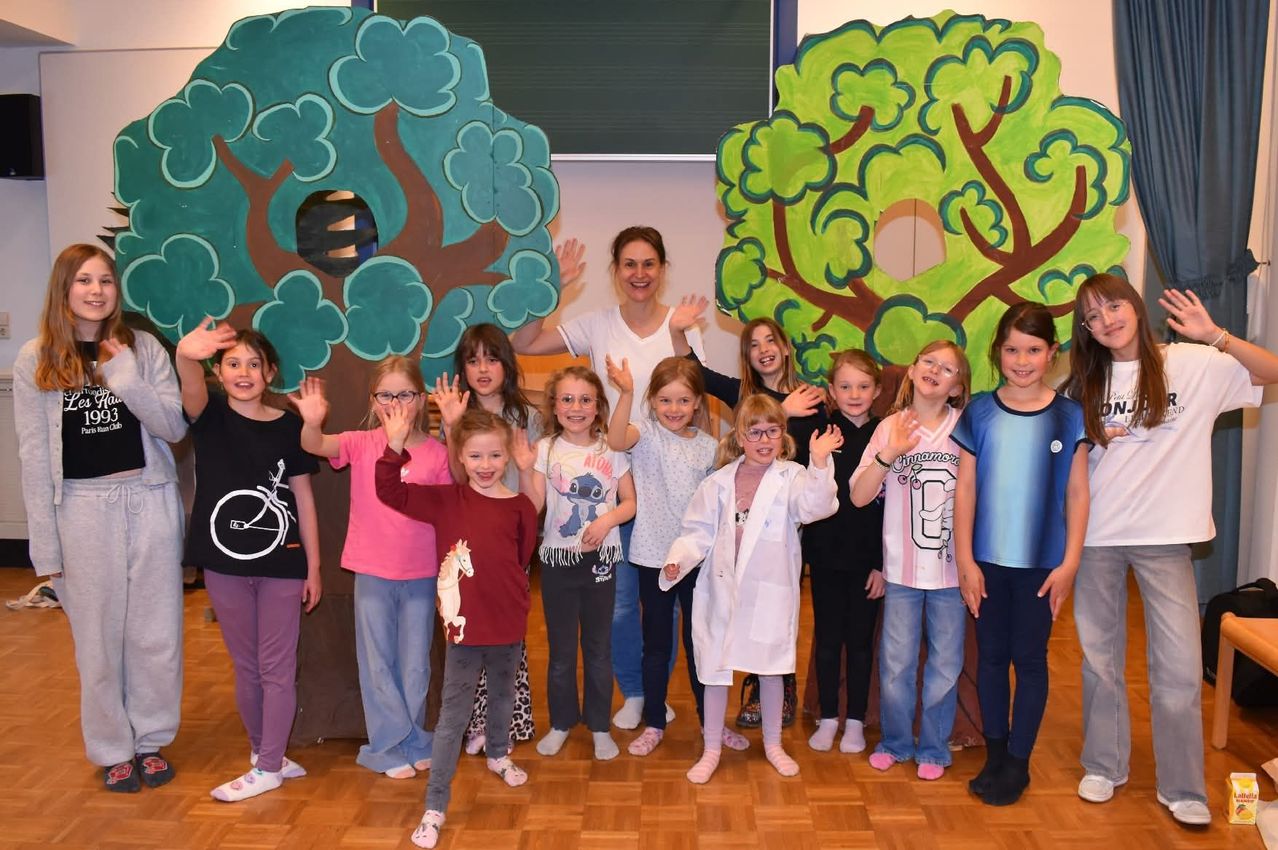 Eine Gruppe von Kindern in einem Klassenzimmer mit einer Lehrerin. Sie stehen hinter Baumausschnitten und machen Handzeichen. Die Lehrerin lächelt und steht in der Mitte.