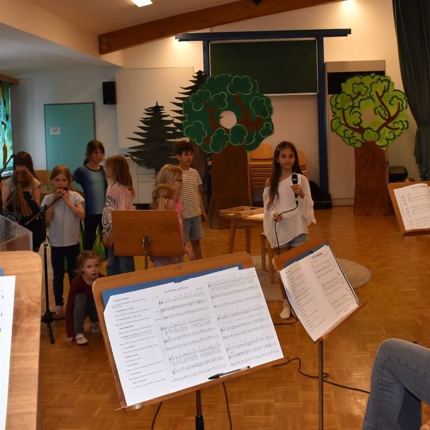 Kinder versammeln sich in einem Raum mit Musikinstrumenten. Ein Mädchen steht vor einem Mikrofon und singt wahrscheinlich, während andere Flöten spielen. Notenblätter stehen vor ihnen auf Ständern.