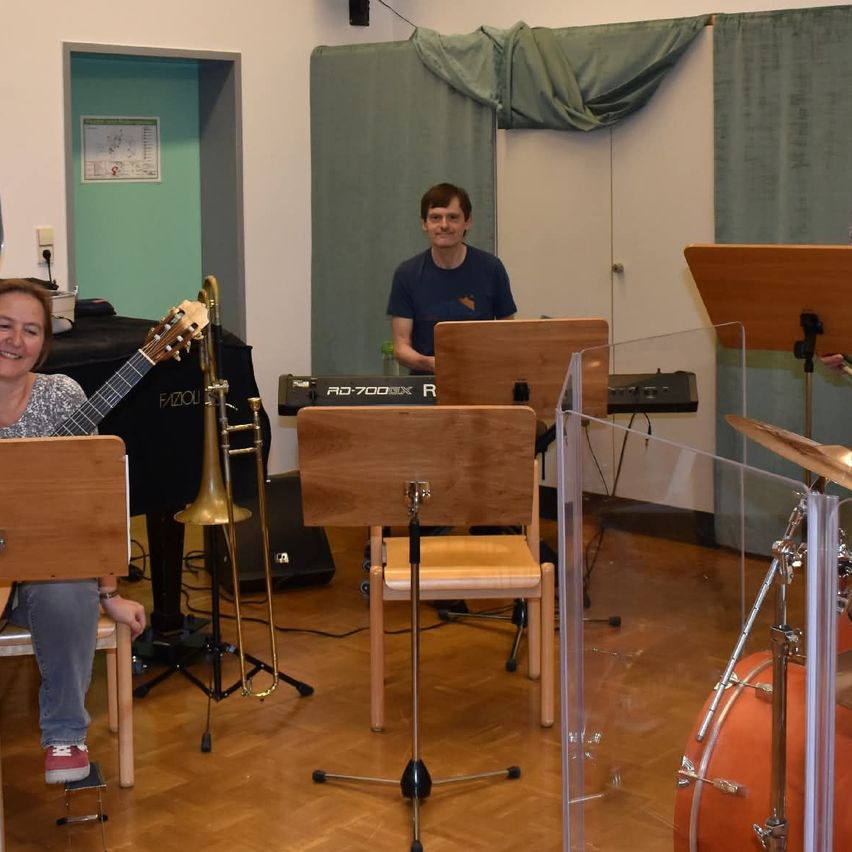 Zwei Personen befinden sich in einem Musikraum. Eine Frau spielt die Gitarre und ein Mann das Klavier. Hinter ihnen steht ein Posaunenständer. Rechts befindet sich ein Schlagzeug.