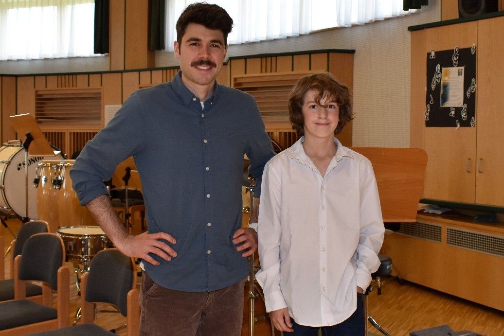 Zwei Personen stehen in einem Musikzimmer, eine lächelt und trägt ein blaues Hemd, die andere ein weißes. Hinter ihnen befinden sich Instrumente, einschließlich Trommeln, und hölzerne Schränke.