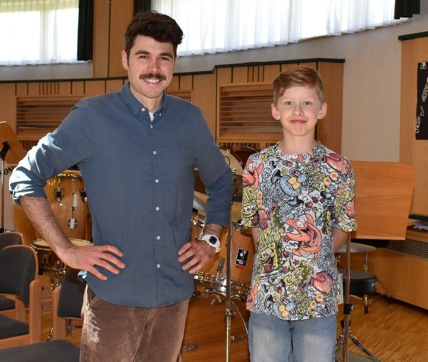Zwei Jungen stehen nebeneinander in einem Raum mit Trommeln und Instrumenten. Der Junge auf der linken Seite trägt ein blaues Hemd und eine Hose, während der Junge auf der rechten Seite ein farbenfrohes Hemd trägt.