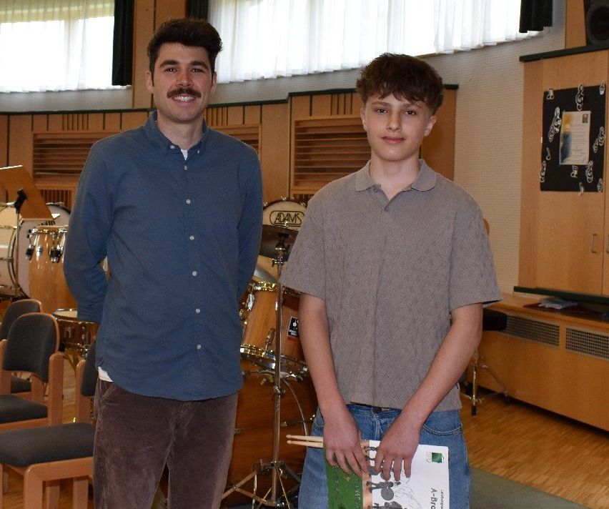 Zwei Jungen stehen in einem Musikzimmer, einer hält ein Buch und Drumsticks, der andere lächelt. Hinter ihnen sind ein Schlagzeug und Holzschränke zu sehen.