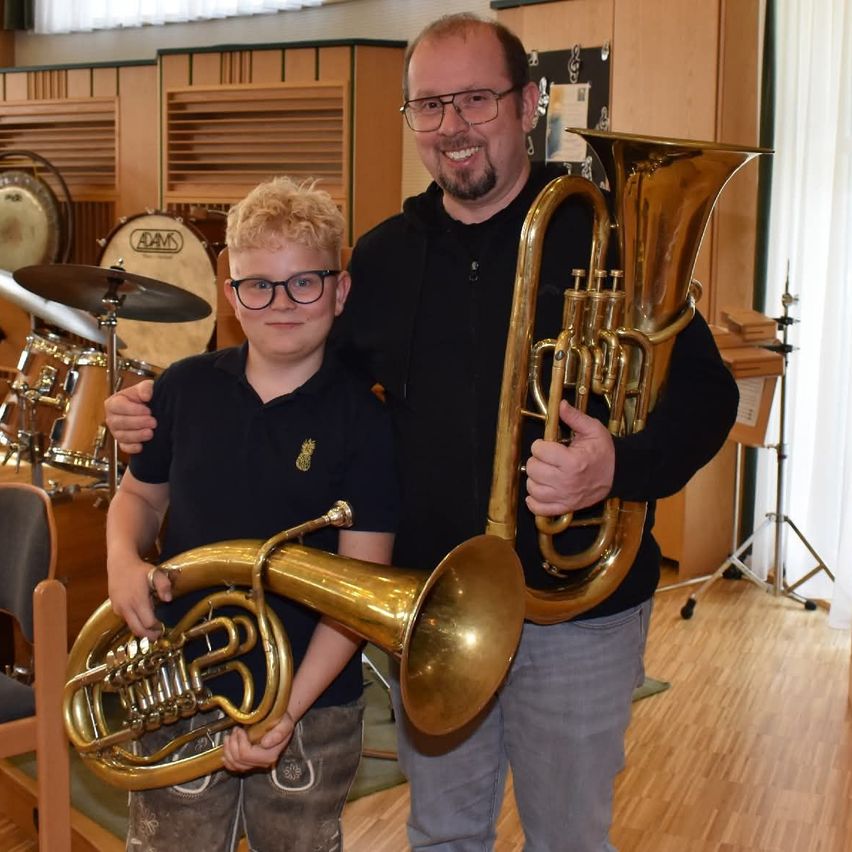 Ein Mann und ein Junge stehen zusammen in einem Musikraum. Der Mann hält ein Blechinstrument und lächelt, während der Junge ein ähnliches Instrument in der Hand hat. Hinter ihnen sind Trommeln und andere Musikinstrumente zu sehen.