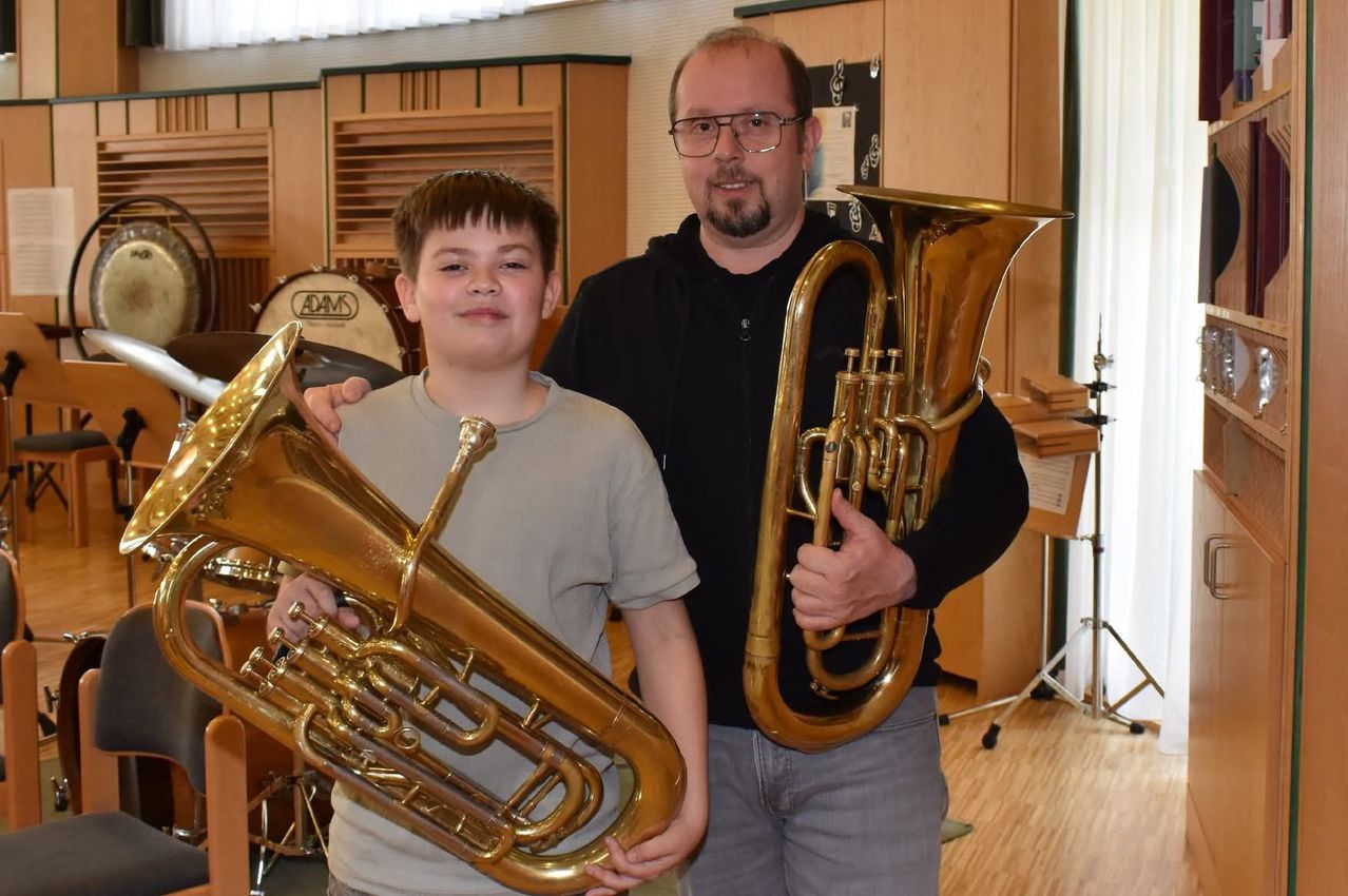 Ein junger Junge mit lächelndem Gesicht und einer Tuba in der Hand steht neben einem Mann mit Brille und Tuba in einem Musikzimmer. Hinter ihnen stehen Musikinstrumente, darunter ein Schlagzeug.
