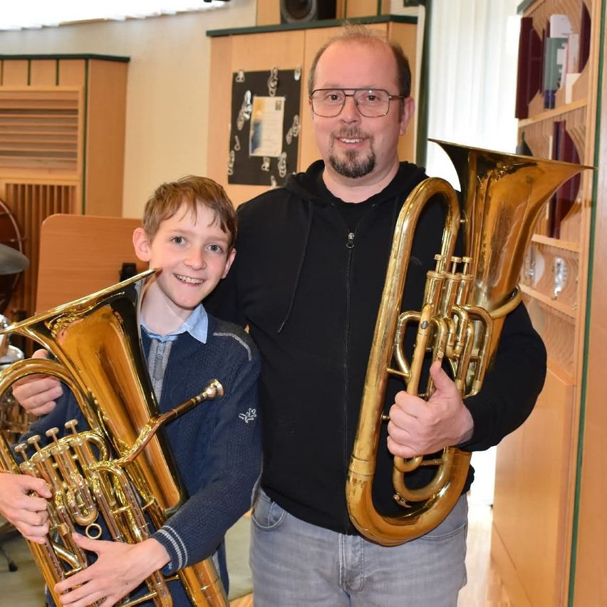 Ein Mann und ein Junge lächeln und halten Blasinstrumente, möglicherweise Tubas, in den Händen. Sie stehen nebeneinander. Dahinter befindet sich ein Holzregal mit verschiedenen Gegenständen. Der Raum ist warm beleuchtet.