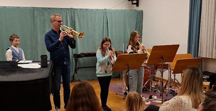 Ein Mann und drei Kinder spielen Trompeten in einem Raum mit Klavier und Publikum. Ein Junge steht neben dem Klavier, während zwei Mädchen mit Notenständern stehen.