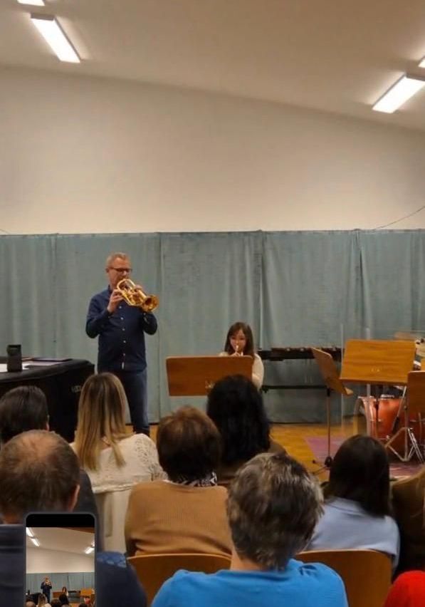 Ein Mann spielt die Trompete vor einem blauen Hintergrund. Eine Frau spielt neben ihm die Flöte. Sie treten vor einem Publikum auf.