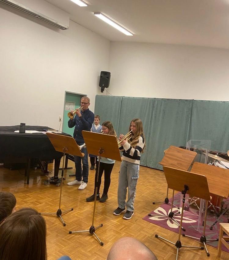 Drei Personen spielen Trompeten in einem Raum mit Holzboden und weißen Wänden. Ein Mann steht neben zwei Mädchen. Ein Klavier und ein Lautsprecher sind im Raum.