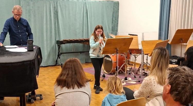 Eine Flötenspielerin steht in einem Raum mit Musikinstrumenten, einem Klavier und einer Menschenmenge. Ein Mann in einem blauen Shirt steht hinter einem Tisch.