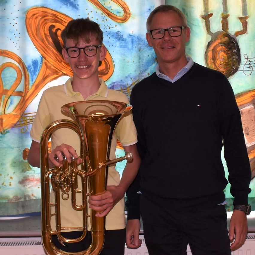 Two men stand next to each other with a large tuba. The young man holds the tuba while the other man smiles at the camera. Behind them is a wall with musical instruments painted on it.