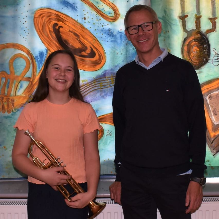 A young girl holds a trumpet, smiling, stands beside a man with glasses and a black sweater, in front of a colorful abstract painting.