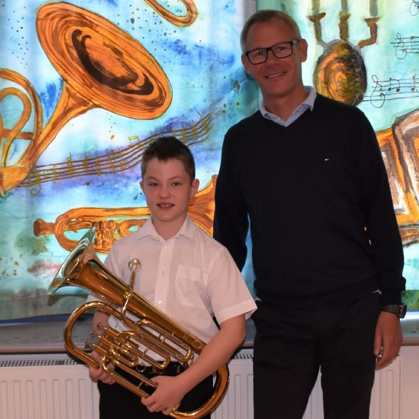 A boy in a white shirt holds a tuba. A man in a black sweater stands next to him. They are in front of a colorful painting with musical instruments.