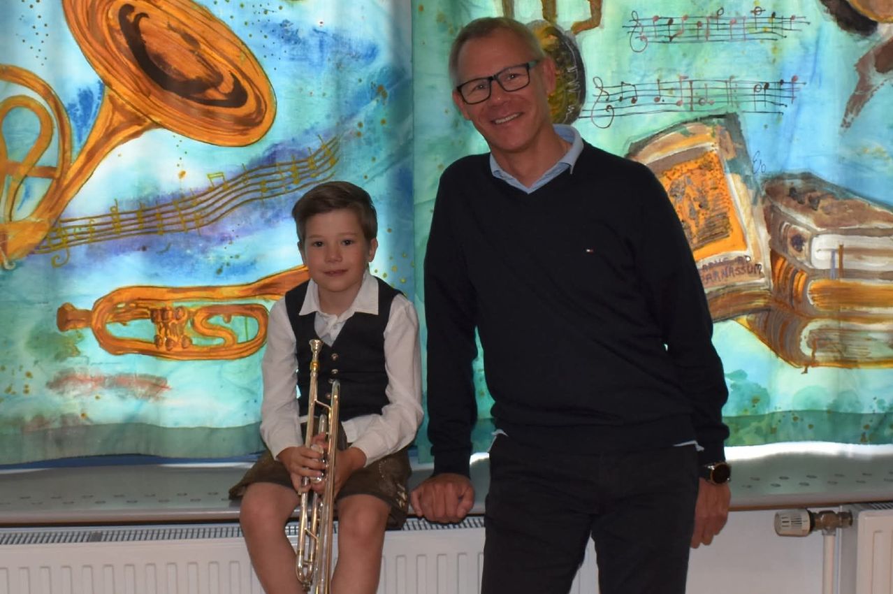 A man and a young boy sit next to each other on a bench. The boy holds a trumpet. They both smile and appear to be posing for a photograph. Behind them, there is a backdrop of musical notes and instruments.
