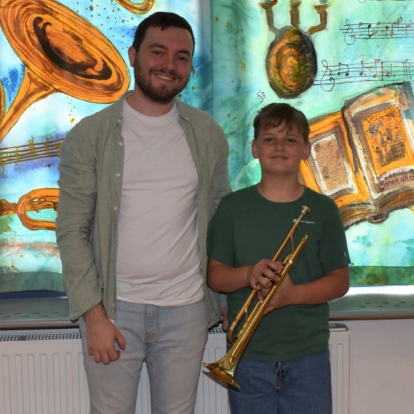 Two individuals stand before a backdrop of musical instruments and notes. The man holds a trumpet and smiles, while the boy holds a trumpet and looks forward.