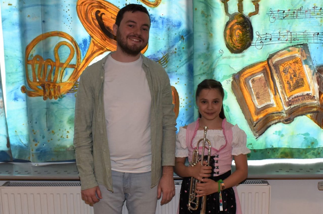 A man in a light green jacket and a young girl in a traditional German dress stand together, smiling for a photo in front of a colorful, musical background.