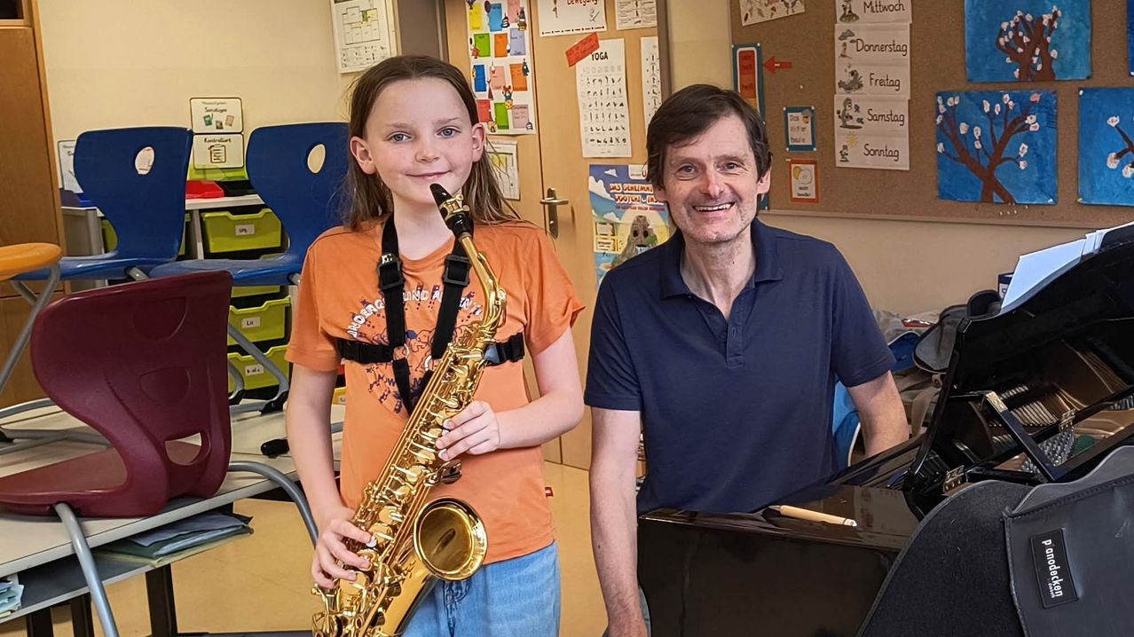 A girl holding a saxophone stands next to a man smiling in a classroom with a piano and bulletin board behind them.