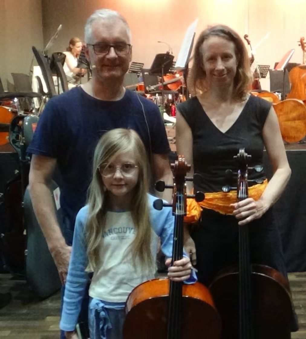 Eine Familie steht lächelnd zusammen in einem Raum voller Musikinstrumente. Das Mädchen hält ein Cello, und die Erwachsenen stehen neben ihr mit Cellos. Sie befinden sich in der Nähe einer Kulisse aus Stühlen, Notenblättern und verschiedenen Instrumenten.