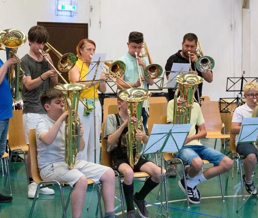 Eine Gruppe von Musikern, sowohl jung als auch erwachsen, spielen Posaunen in einem Raum mit grünem Boden. Einige sitzen, andere stehen, alle schauen in ihre Notenblätter.
