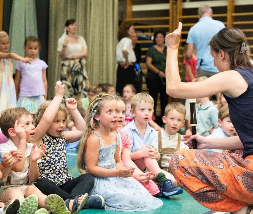 Eine Frau unterrichtet eine Gruppe von Kindern, die auf dem Boden sitzen, und hält eine Flöte. Sie lauschen aufmerksam.