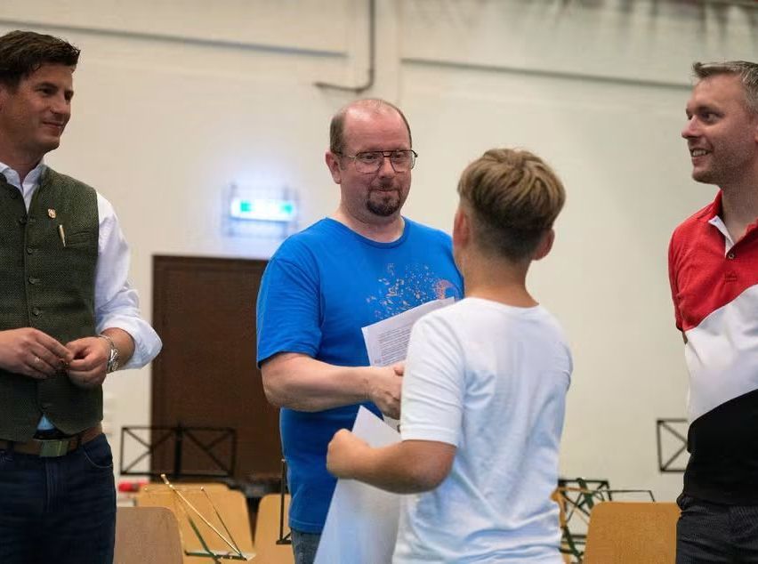 Ein Mann in einem blauen T-Shirt überreicht einem jungen Jungen eine Urkunde. Sie werden von drei anderen Männern umringt, einer in einem rot-weißen Shirt. Die Umgebung scheint ein Probensaal mit Stühlen und Musikinstrumenten zu sein.