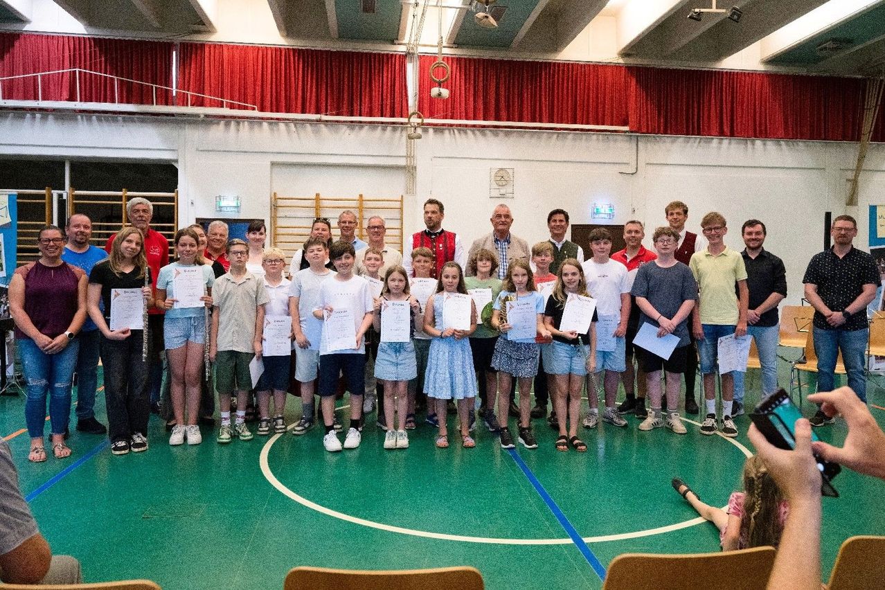 Eine Gruppe von Kindern und Erwachsenen posiert für ein Foto in einer Turnhalle mit Urkunden in der Hand. Sie stehen auf einem grünen Mattenboden, und eine Person liegt auf dem Boden. Hinter ihnen hängt ein roter Vorhang.