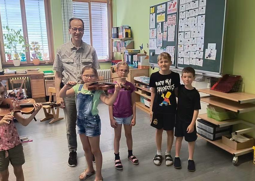 Ein Mann mit Brille steht in einem Klassenzimmer mit vier Kindern. Sie halten Geigen. Ein Regal und eine Pinnwand sind hinter ihnen.