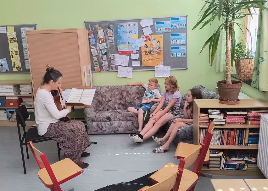 Eine Frau spielt Geige, während sie in einem Klassenzimmer sitzt. Drei Kinder sitzen auf einer Couch hinter ihr, und Bücher und Pflanzen sind im Raum verteilt.