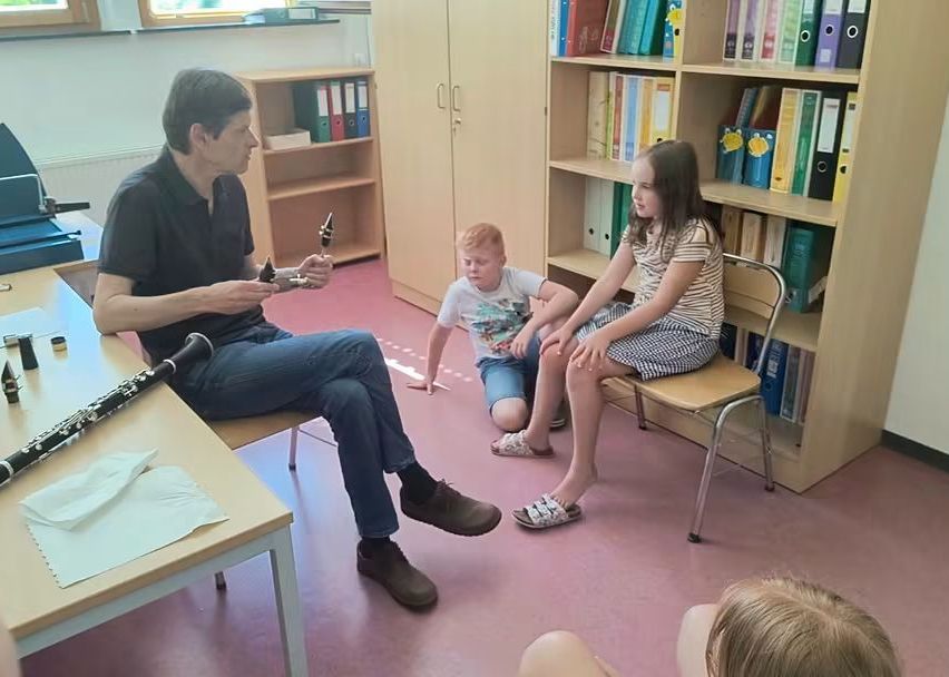 Ein Mann spielt eine Klarinette, sitzt auf einem Stuhl in einem Klassenzimmer mit drei Kindern, zwei sitzen auf Stühlen und eines kniet auf dem Boden, mit Bücherregalen dahinter.