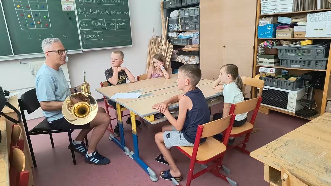Ein Musiklehrer spielt ein Tenorhorn für eine Gruppe von Kindern, die an Tischen sitzen. Sie wirken aufmerksam, und ein Tafel mit Schriftzeichen ist im Hintergrund zu sehen.