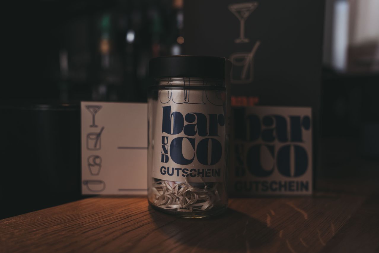 A glass jar with a black lid filled with strips of paper, labeled 'bar und co Gutschein,' sits on a wooden table with a blurred background.